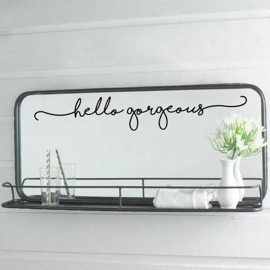Sticker met de tekst “hello gorgeous” op een spiegel met een zwarte plank, een vaas met bloemen en glazen