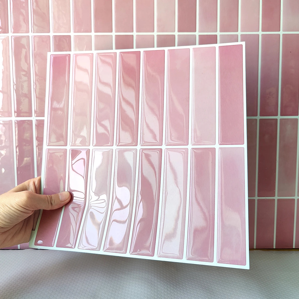 Hand holding a glossy pink tile sticker sheet in front of a tiled wall