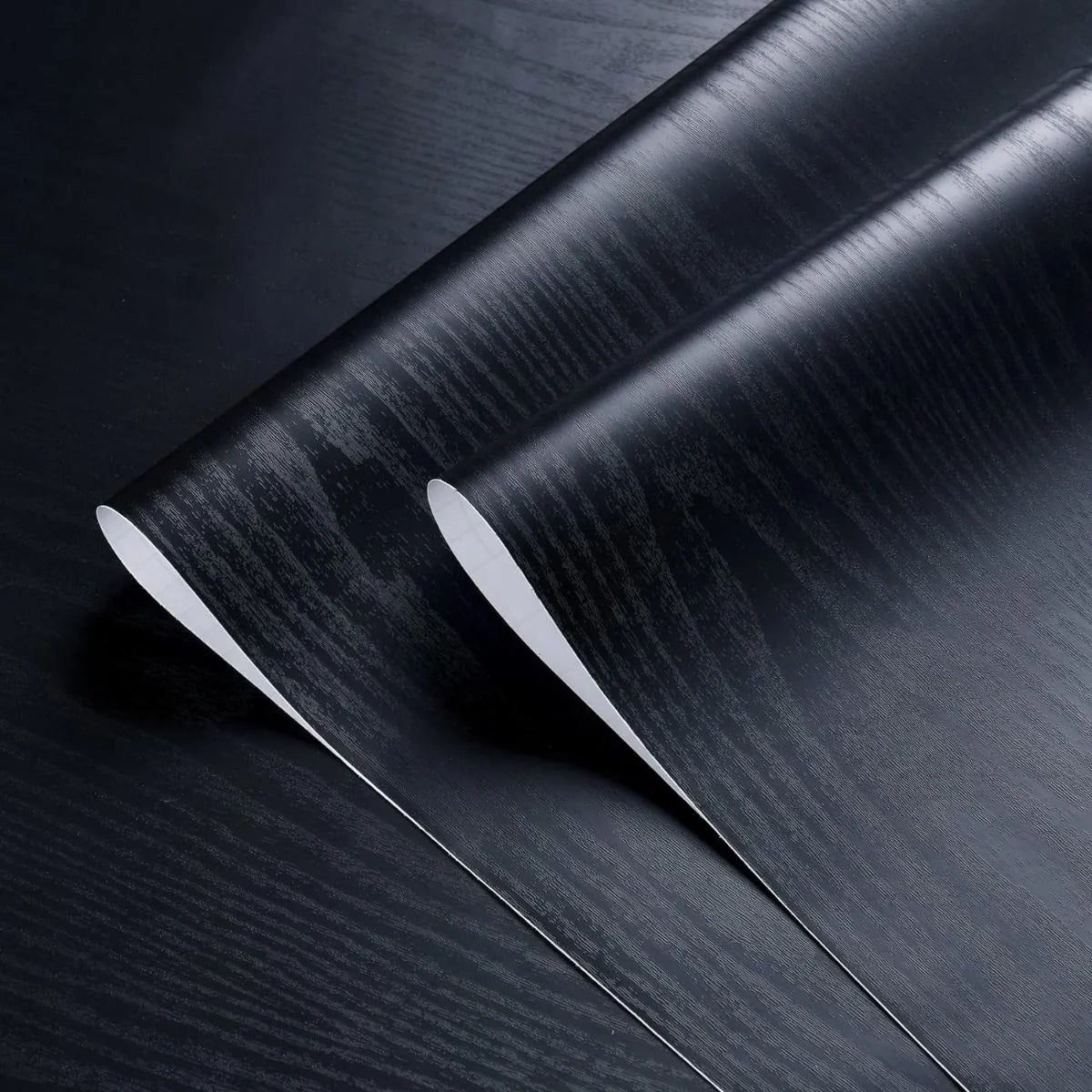 Two rolls of black wood effect vinyl unrolled: embossed grain and white paper backing