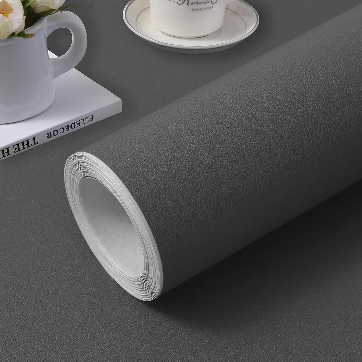 Rolle mattes dunkelgraues selbstklebendes Papier auf grauer Fläche, mit einem Buch und einer Tasse