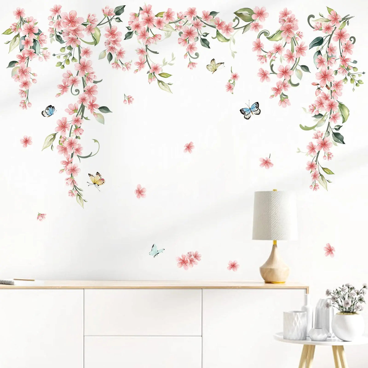 Roze klimbloemen-stickers met vlinders op een witte muur boven een laag kastje met een lamp